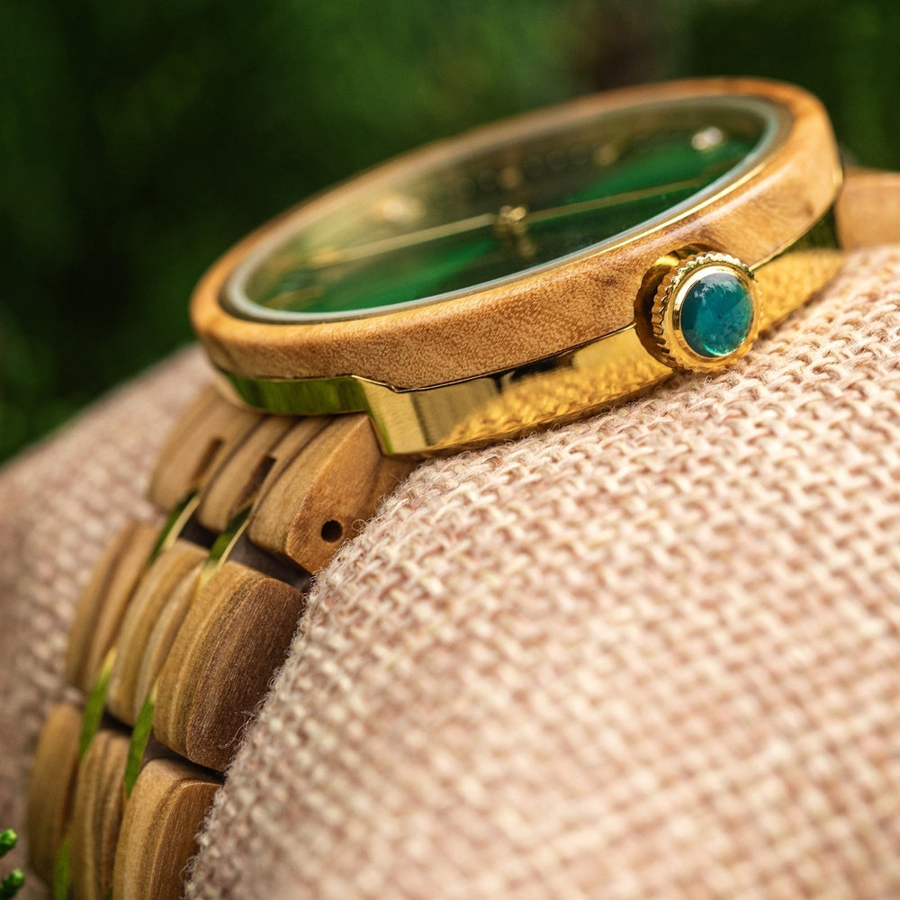 Close-up of a Mokozai wristwatch showcasing the side view of the wooden case and gold-tone crown set with a vibrant emerald gem. The wooden bracelet features gold accents, adding a touch of luxury to the natural wood finish, with the watch resting on a textured cushion.