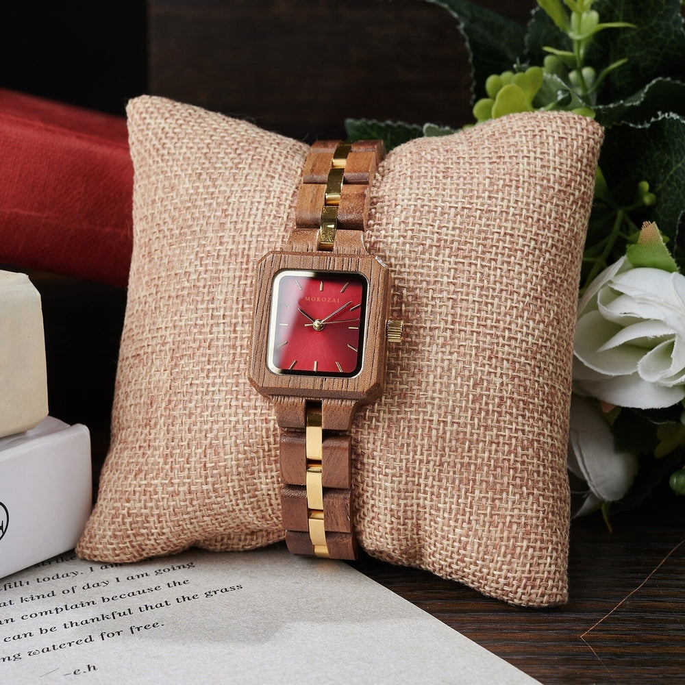 Mokozai square wooden wristwatch with a vibrant red dial and gold accents, elegantly displayed on a beige cushion, surrounded by greenery and books for a natural, luxurious setting.