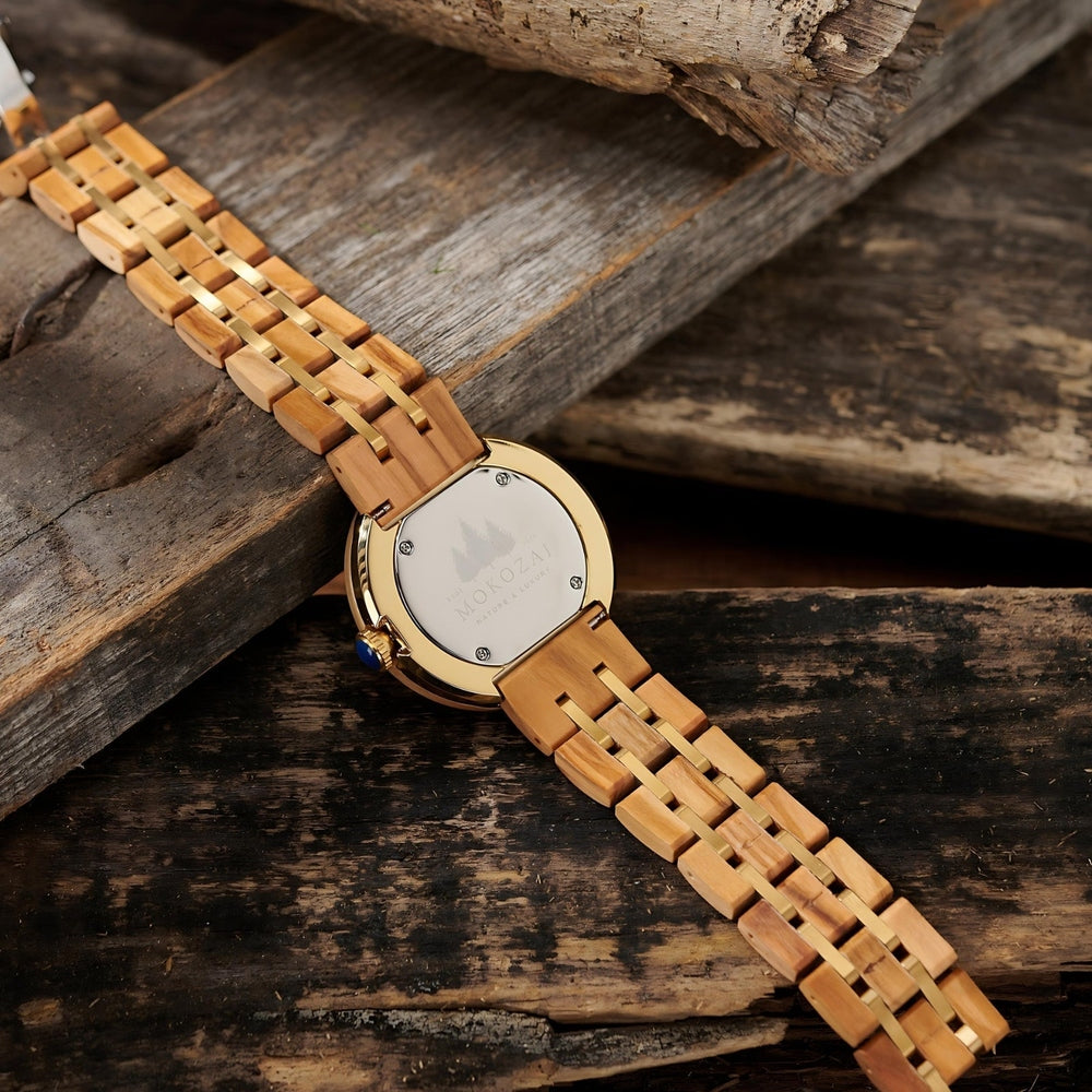 Back view of a Mokozai wooden wristwatch, highlighting the engraved case back with a nature-themed design. The golden accents on the wood bracelet complement the natural wood grains, resting against a rustic wooden surface.