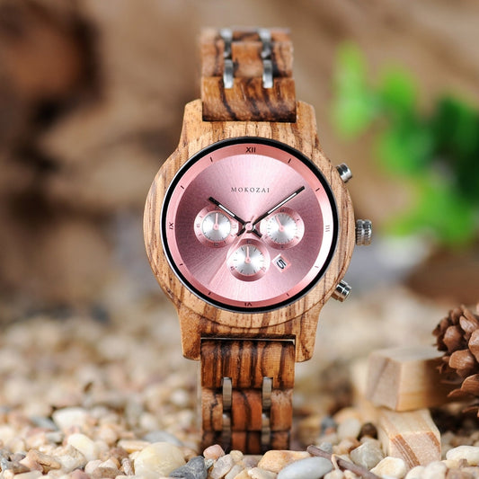 Stylish wooden wristwatch featuring a pink dial and silver sub-dials, paired with a natural wood grain strap, displayed on a bed of pebbles with a natural green background.