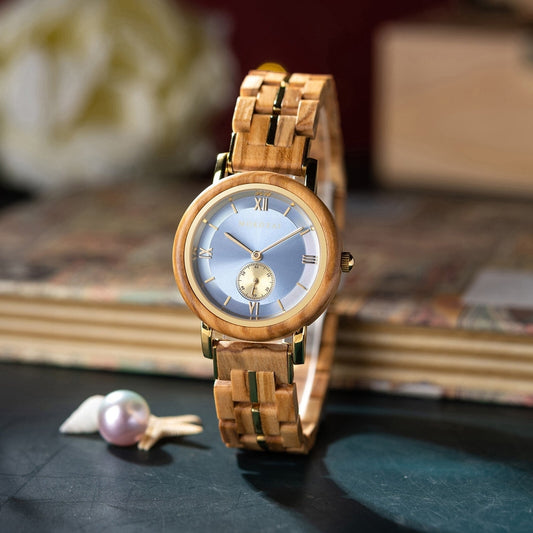 Elegant Mokozai wooden wristwatch with a polished light wood band, gold accents, and a soft blue dial featuring Roman numerals and a small subdial, placed on a dark surface next to a pearl and seashell.