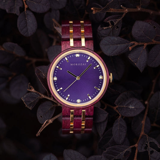 A Mokozai wooden wristwatch with a striking purple dial, gold hands, and crystal hour markers, set against a deep red and gold wooden band, displayed amid dark foliage.