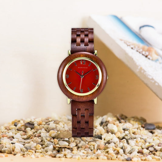 Mokozai wooden watch with a vibrant red dial, gold accents, and a smooth wooden bezel. The watch is elegantly displayed on a bed of natural pebbles, highlighting its earthy and luxurious appeal. The wooden bracelet enhances the rustic yet sophisticated design, perfect for fashion-forward individuals.